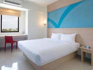 Minimalist hotel room with a clean white bed, vibrant blue accent wall, and a simple red chair, presenting a modern and sleek accommodation space.