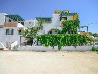 Prédio branco com trepadeiras verdes, varandas e uma placa com a palavra "Domingo" no telhado, sob um céu azul claro.