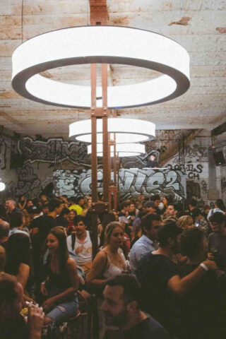 A bustling nightlife scene at La Calle Bar, with a large crowd mingling under unique circular light fixtures, against a backdrop of graffiti-adorned walls