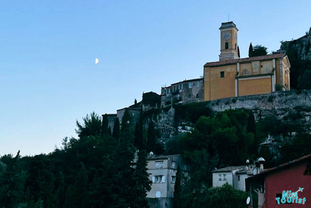 Twilight view of the hilltop village of Èze, France, with buildings perched on a rocky outcrop and the crescent moon above
