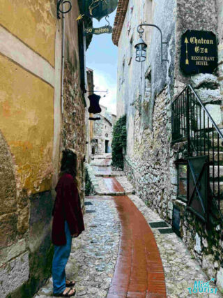 The writer of the post contemplates the quaint, stone-paved streets of Èze, France, lined with traditional buildings and signs pointing to a local hotel and restaurant
