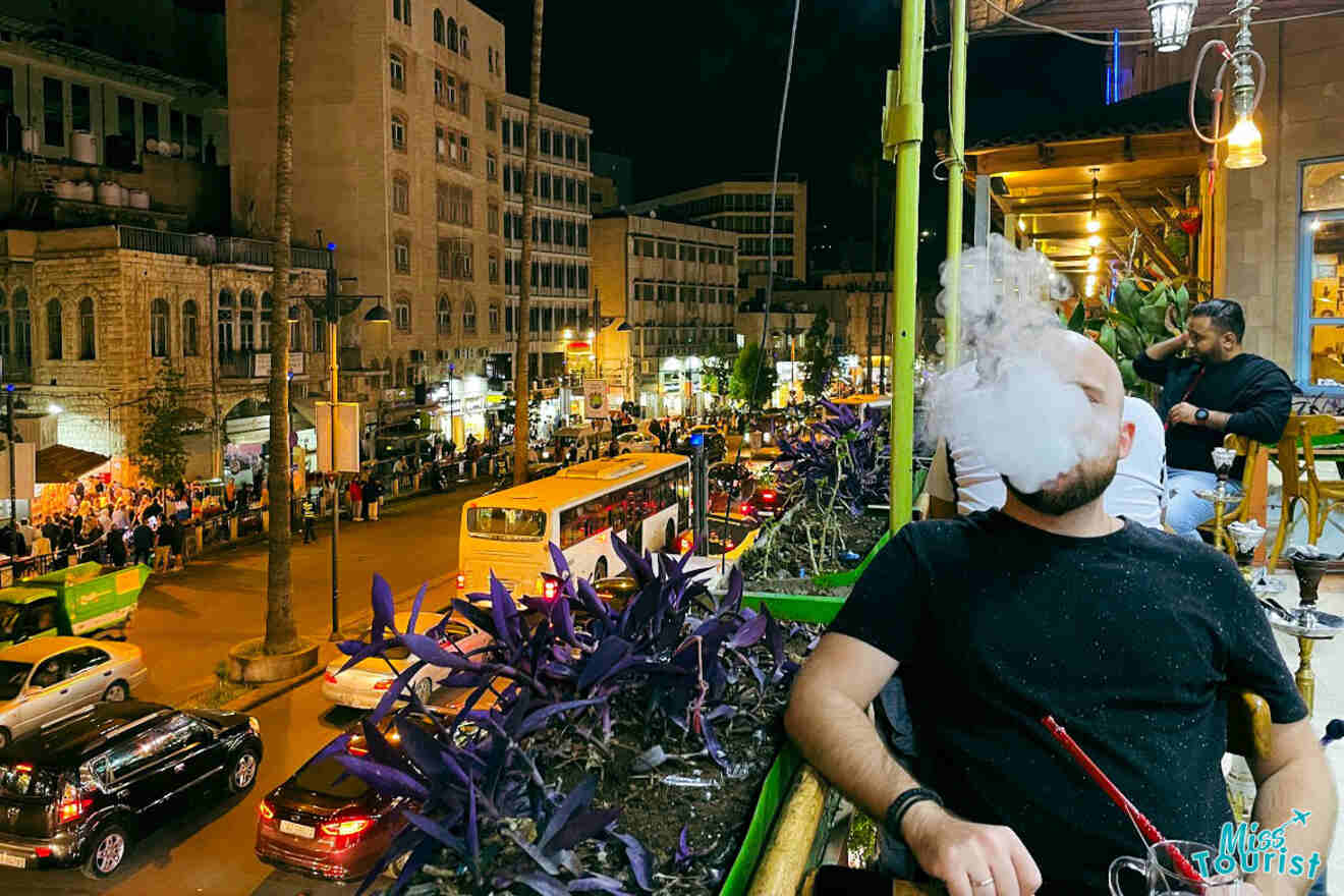 A man enjoying shisha at a local café in Amman, Jordan, with the busy street and lit shops in the background