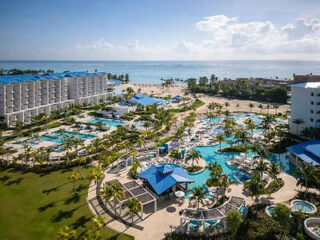 Aerial view of a beachfront resort featuring multiple pools, palm trees, white buildings with blue roofs, and the ocean in the background.