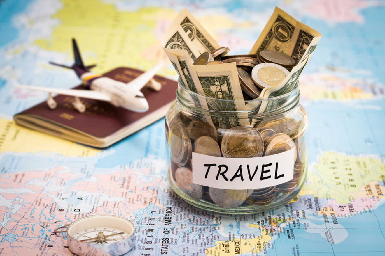 A clear glass jar labeled 'TRAVEL' filled with coins and paper currency sits atop a colorful map, flanked by a passport with a small airplane model on top and a compass