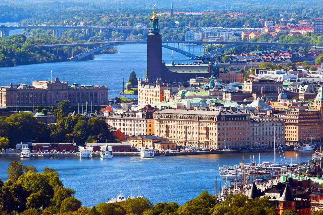 Panoramic view of Stockholm, showcasing the iconic City Hall with its tall tower and golden Three Crowns, the Royal Palace, and surrounding historic buildings along the waterfront, with bridges connecting to lush green islands in the background.