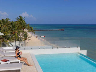 A person in a swimsuit sits beside an infinity pool overlooking a sandy beach and the ocean under a clear blue sky.