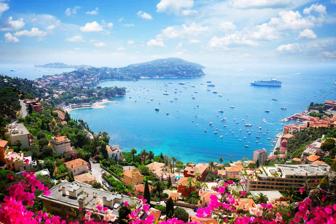 A panoramic view of the French Riviera, showcasing the azure waters of the Mediterranean Sea dotted with numerous boats, the lush greenery of the hills, and the terracotta rooftops of coastal buildings, framed by vibrant pink flowers in the foreground.
