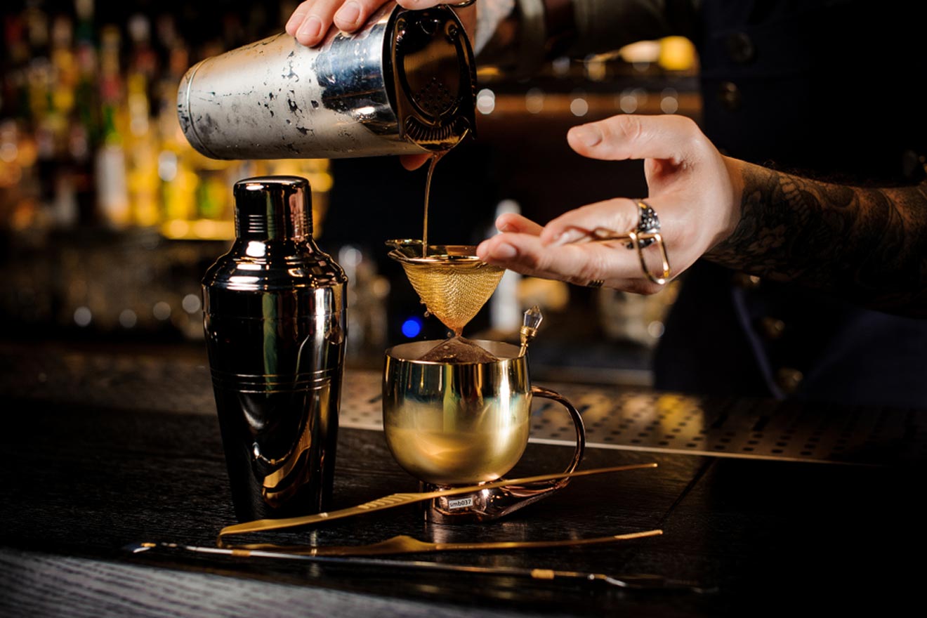A bartender with tattooed arms strains a cocktail through a fine mesh sieve into a golden cup, on a bar counter surrounded by various cocktail making tools in a buenos aires speakeasy.