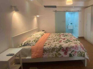 A neatly made bed with floral bedding and an orange blanket in a modern bedroom; sliding glass door leads to a brightly lit bathroom.