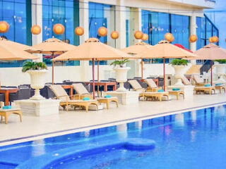 Outdoor pool at The Kingsbury Colombo, lined with cream parasols, sun loungers and urn planters against the hotel's glass facade.