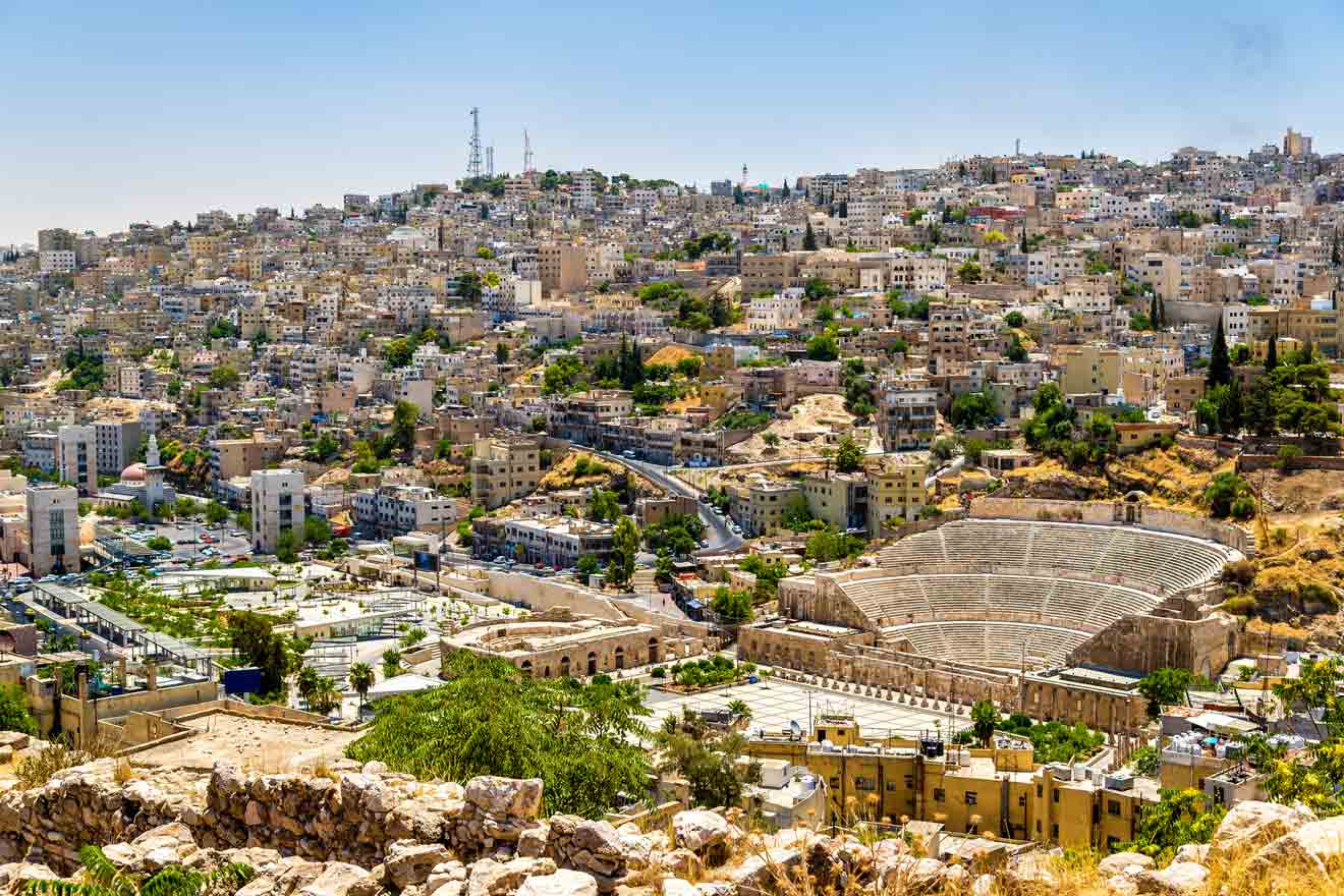 Panoramic view of Amman, Jordan showcasing the densely packed buildings in various shades of white and beige, with the ancient Roman theater in the foreground. The city's hills create a staggered skyline under a clear blue sky