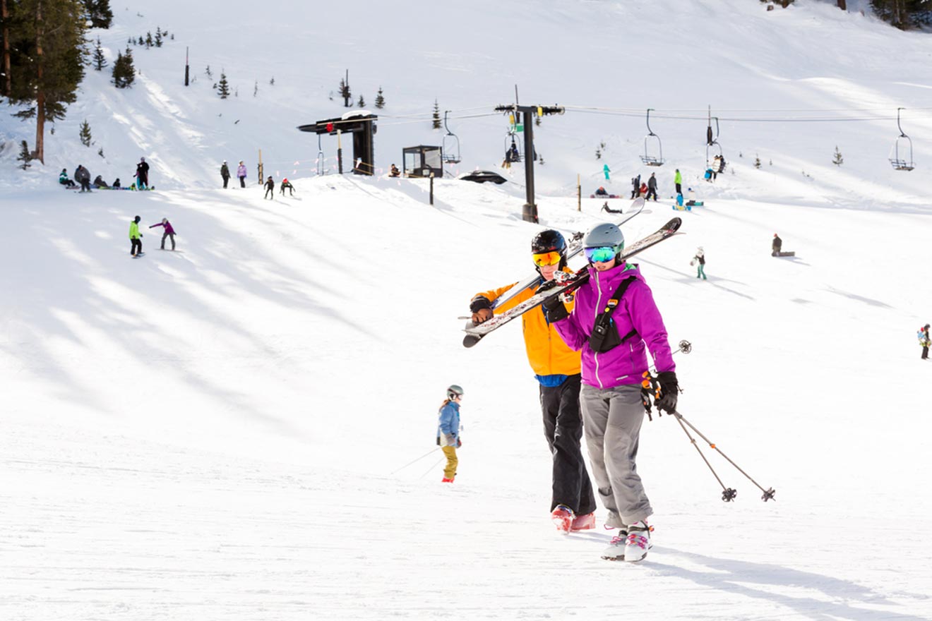 Two skiers carrying their skis walk across a snowy slope at a ski resort, with other skiers and chairlifts visible in the background, on a bright sunny day in Vail, Colorado