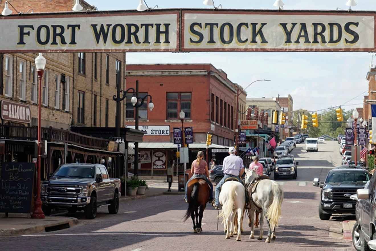 Visitors on horseback explore the historic Fort Worth Stockyards under a large sign that reads 'Fort Worth Stock Yards', with classic brick buildings, various storefronts, and parked cars lining the street.