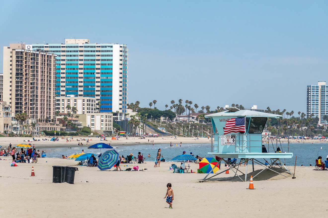 A bustling beach scene with a distinctive light blue lifeguard tower flying the American flag in the foreground. Beachgoers relax under colorful umbrellas with a backdrop of tall hotels and palm trees in Long Beach.