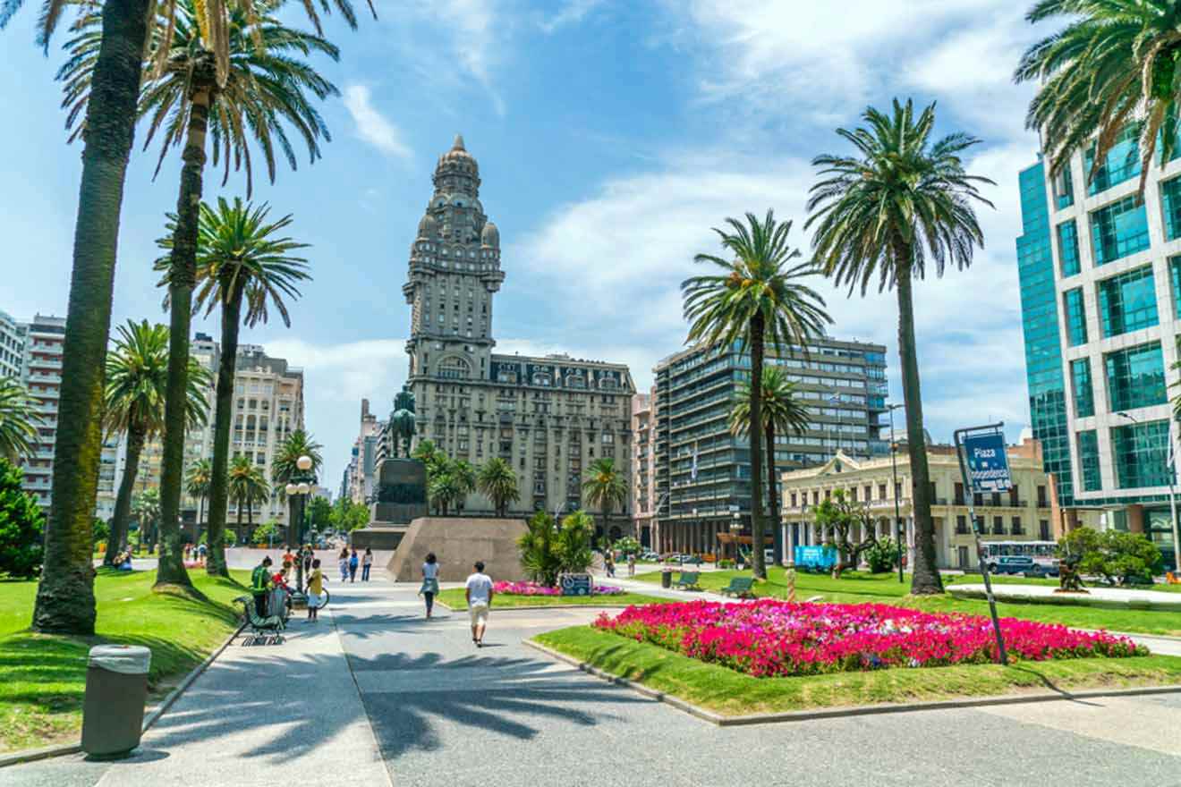 A sunny day in a bustling Montevideo square with tall palm trees, people walking, and a prominent historic building with a tower in the background, surrounded by modern structures and vibrant flowerbeds.
