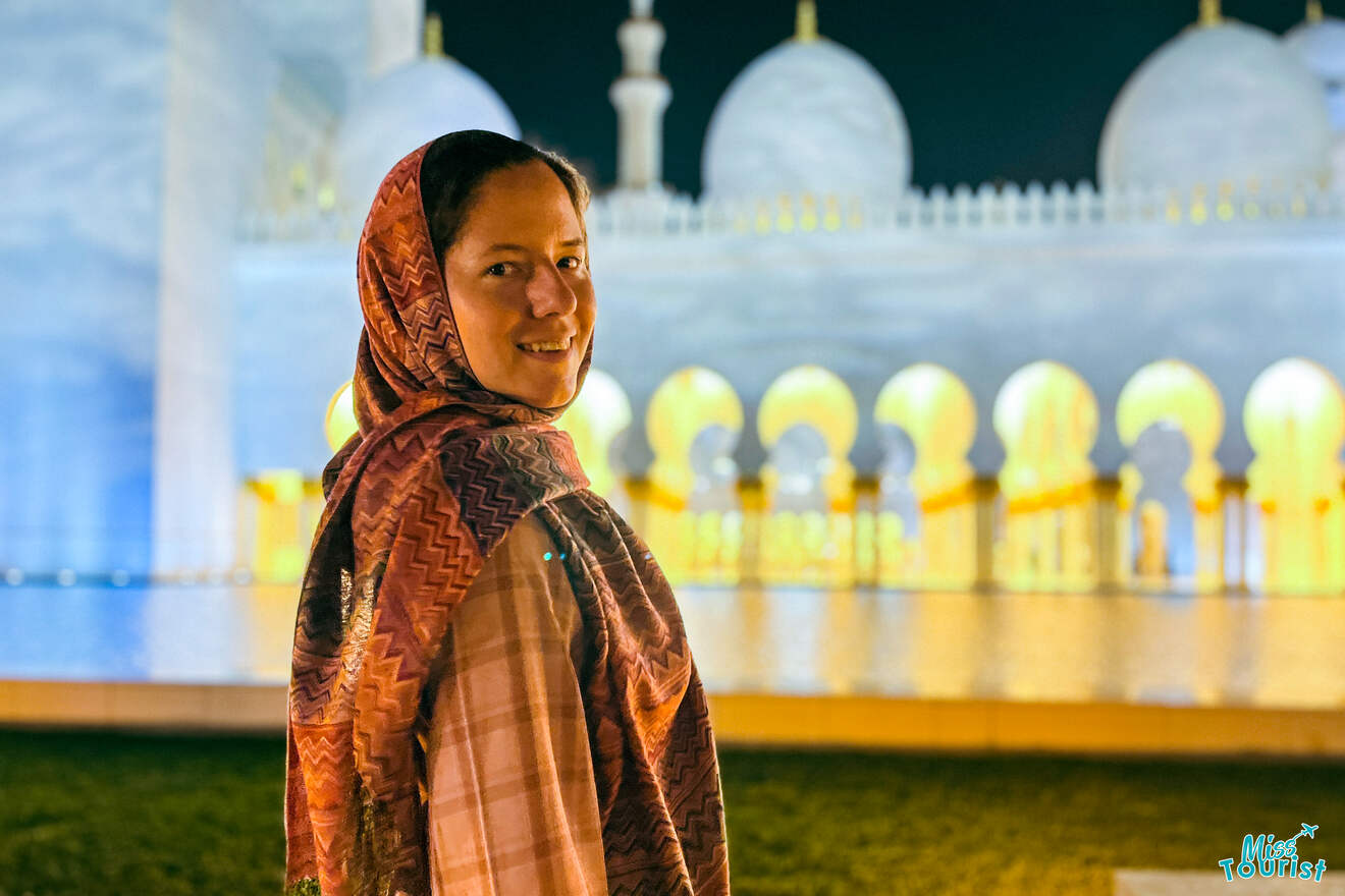 Writer of the post wearing a patterned headscarf stands in the foreground, smiling gently, with the brilliantly lit Sheikh Zayed Grand Mosque forming a luminous backdrop at night,