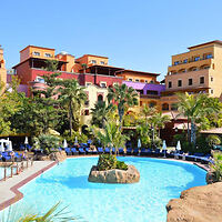 Bunte, mehrstöckige Hotelgebäude umgeben einen Swimmingpool mit Liegestühlen, Sonnenschirmen, Palmen und einer kleinen Felseninsel in der Mitte.
