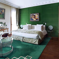 A chic bedroom with emerald green walls and carpet, featuring a large bed with patterned coverlet, white linens, and a painting above the headboard.