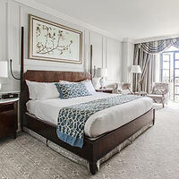 Luxurious bedroom in The Charleston Place hotel featuring a king-sized bed with a dark wood headboard, white and patterned bedding, elegant wall art, and access to a balcony with a view