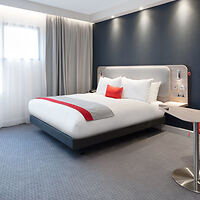 Contemporary and minimalist hotel room with a king-sized bed accented with a red throw pillow, flanked by sheer curtains and dark blue walls.