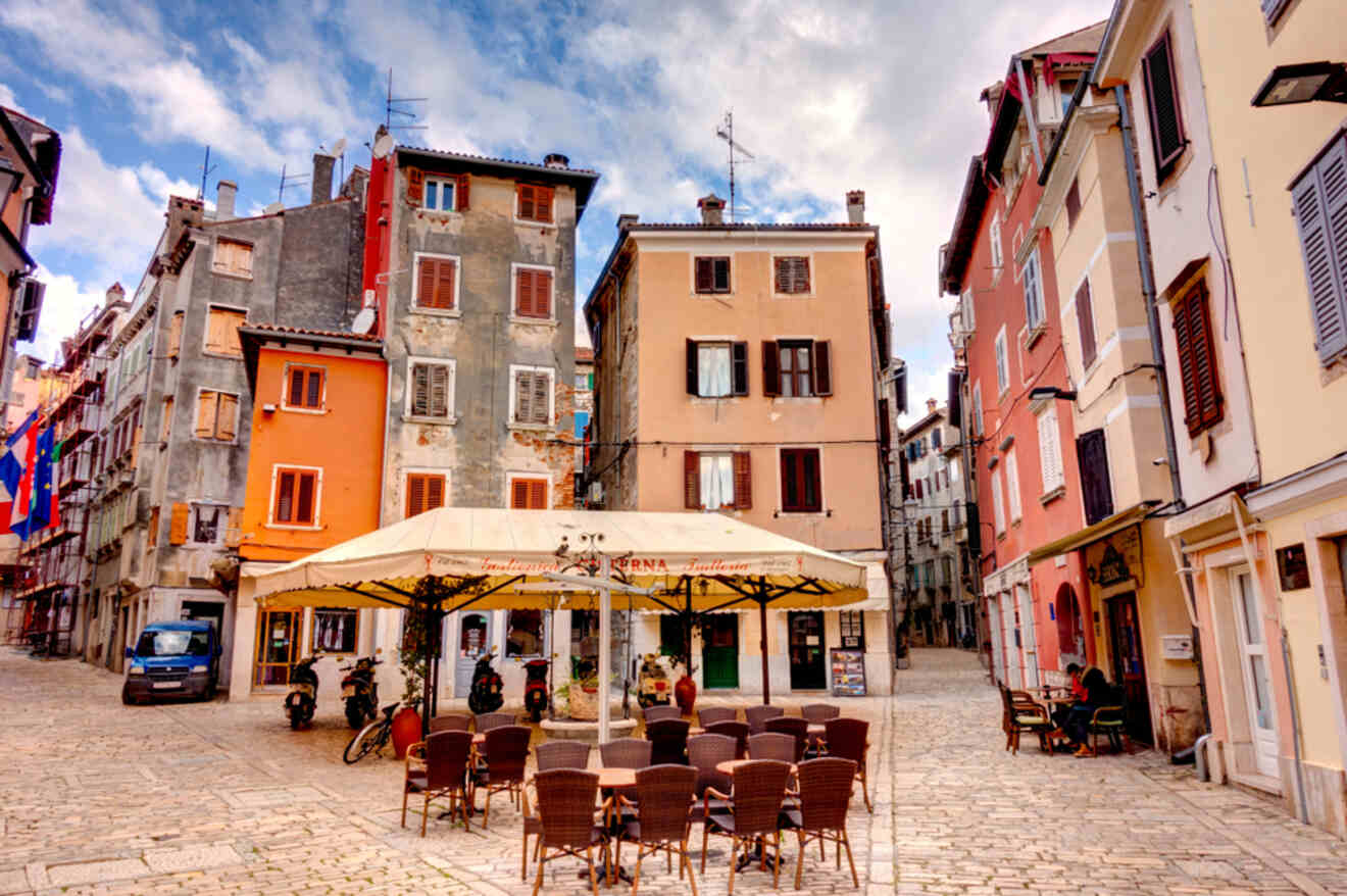 Quaint outdoor cafe nestled among colorful, historic buildings on a cobblestone street in Rovinj, under a clear blue sky with fluffy clouds