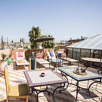 Sun-drenched rooftop terrace with mosaic-tiled tables, colorful chairs, and lounge areas, offering a view of the Marrakech skyline