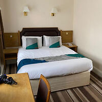 Hotel room with a double bed, accentuated with teal pillows, a wooden desk