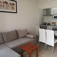 A small living room with a grey sectional sofa, a wooden side table, a dining table with four white chairs, and a compact kitchen area in the background.
