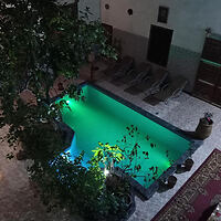 Evening view of a serene riad courtyard with a glowing turquoise pool surrounded by green foliage and traditional Moroccan tiles