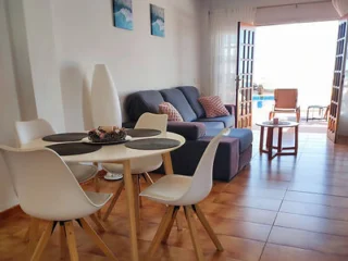 A modern living-dining area with a round table and four white chairs, a gray sofa, and open doors leading to a bright terrace.