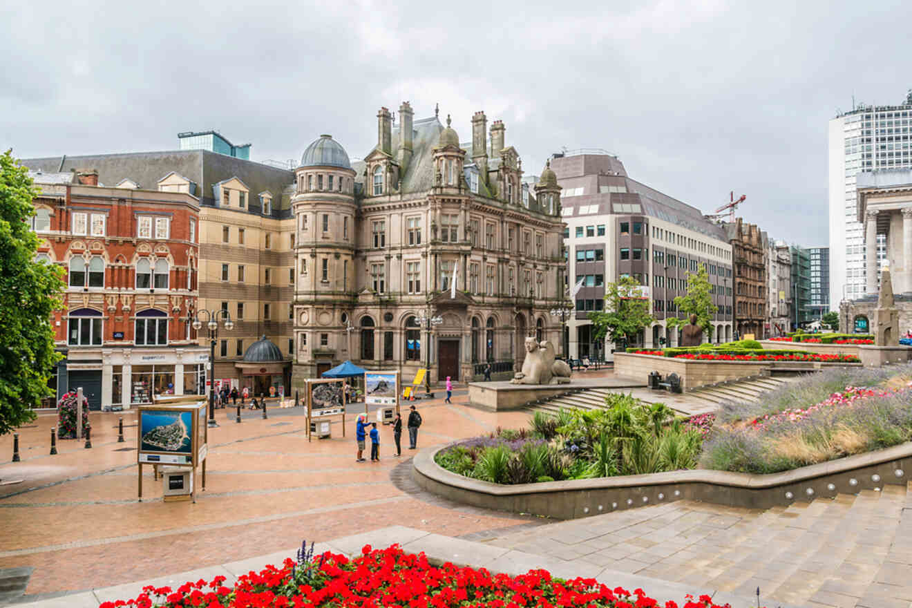 Cityscape of Birmingham City Centre with a mix of historic and modern architecture, featuring prominent buildings, pedestrian walkways, and vibrant red flowers in the foreground