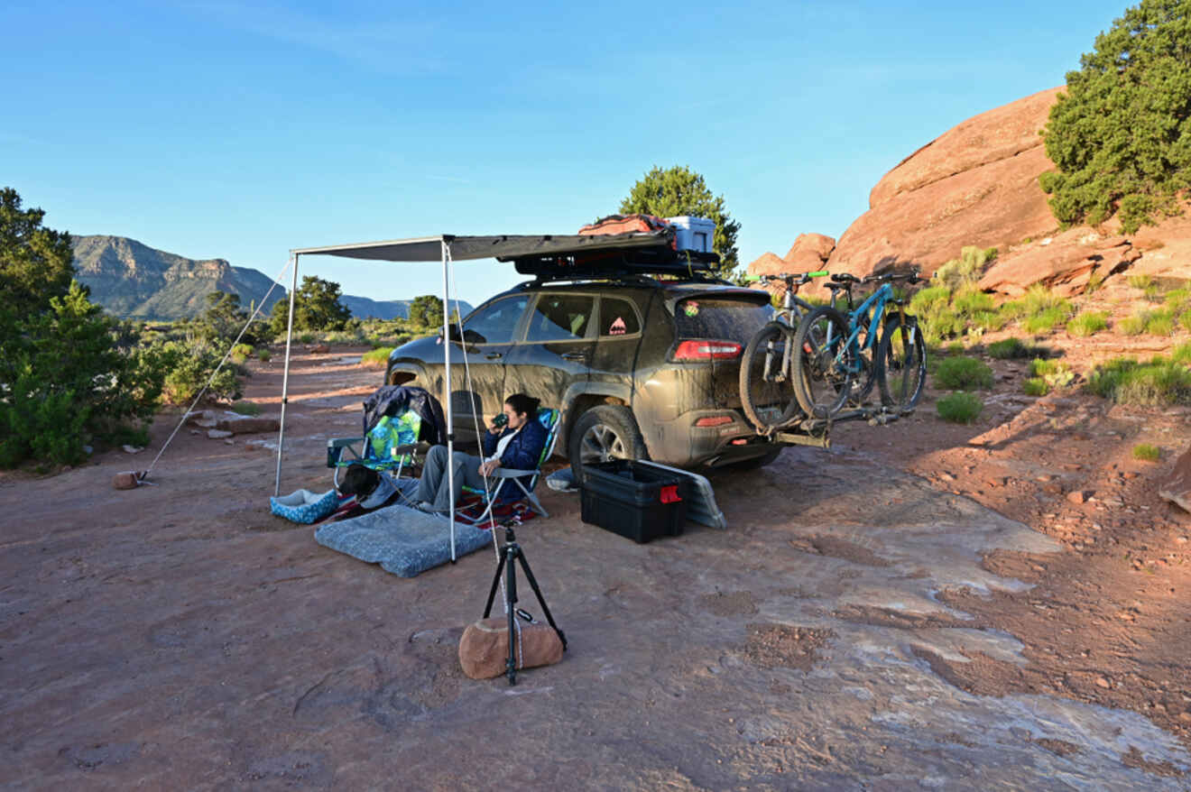 Campsite setup with a car equipped with a roof rack and bicycles attached, a portable canopy, and two people lounging in camping chairs amidst scenic red rock formations