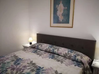 A bedroom with a double bed, floral bedspread, two bedside tables with lamps, and a framed painting on the wall above the headboard.