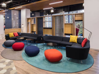 Modern hotel lounge with a teal circular rug, modular black sofa sections adorned with colorful pillows, and circular red, navy, and teal ottomans on a patterned floor