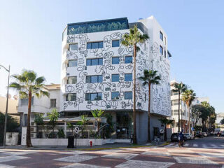 Facade of JM Suites Hotel, featuring a modern design with white walls adorned with bold black artistic scribbles and palm trees flanking the sides, set against a clear blue sky