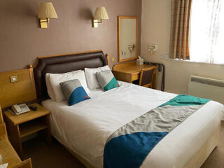 A standard hotel bedroom with a double bed featuring teal and white bedding, wood furnishings, and soft wall lighting