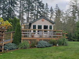 Charming cottage with a spacious wooden deck surrounded by lush landscaping and towering evergreens