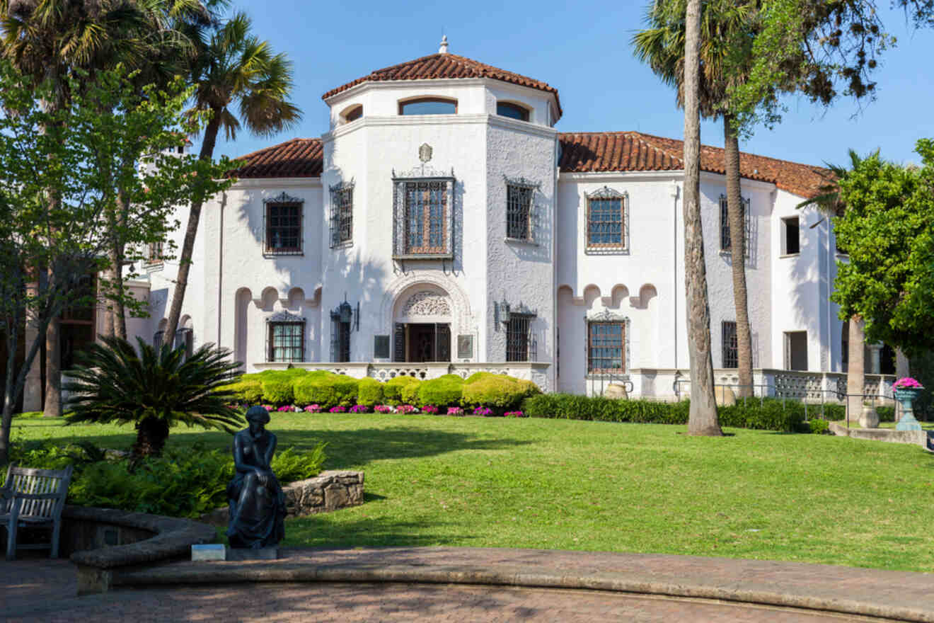 Mediterranean-style building of the San Atonio Museum of Art with a terracotta roof, ornate windows, and a manicured garden featuring a bronze statue.