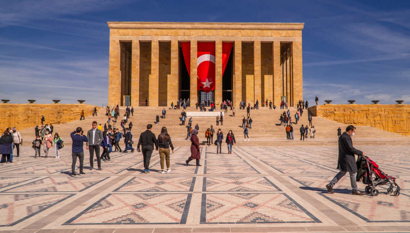 A wide view of Anıtkabir, the mausoleum of Mustafa Kemal Atatürk in Ankara, with a large Turkish flag hanging between two pillars and crowds of visitors on its expansive grounds