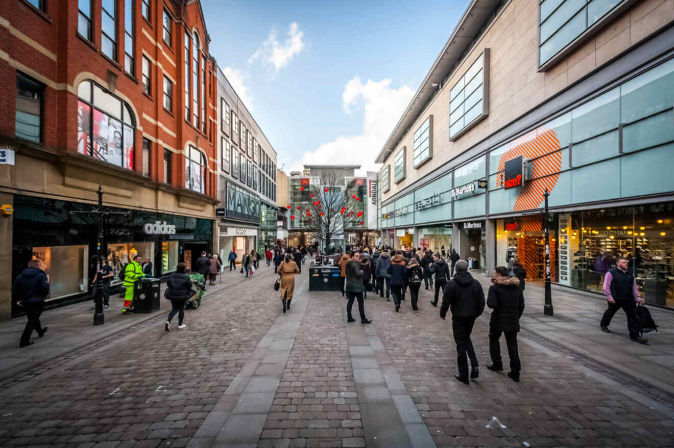 A bustling Market Street in Manchester with pedestrians shopping and walking past high street stores like Adidas and Mango under a clear blue sky