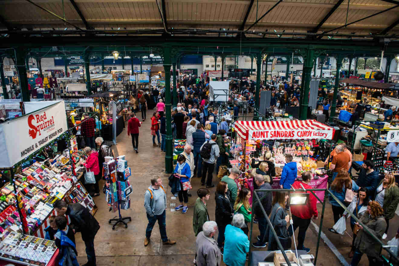 Busy interior scene of St. George's Market in Belfast, with vendors selling diverse goods, from mobile accessories to local candy, to a crowd of shoppers.