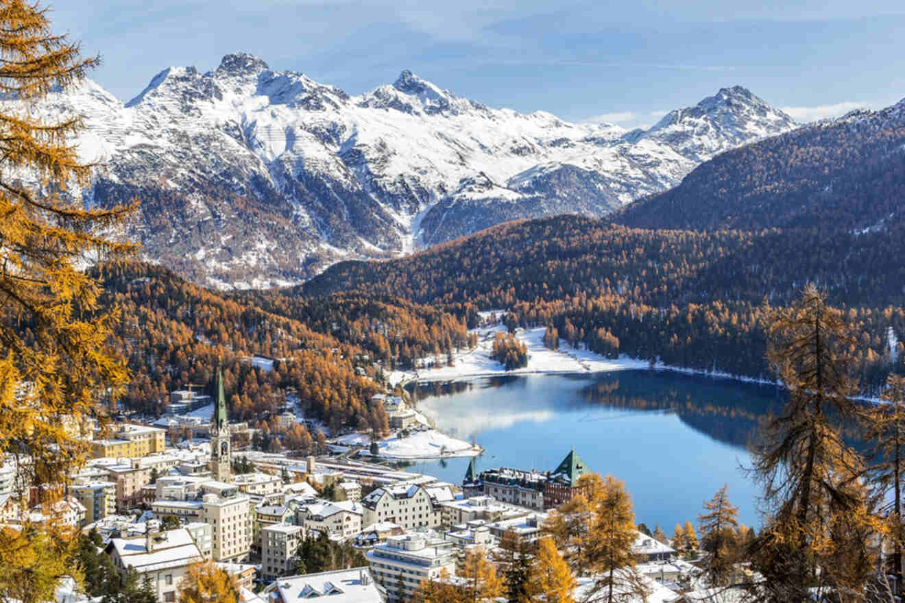 Panoramic view of St. Moritz Dorf in Switzerland with luxury hotels clustered around the frozen Lake St. Moritz, set against a backdrop of snow-covered peaks and autumnal larch trees under a clear blue sky