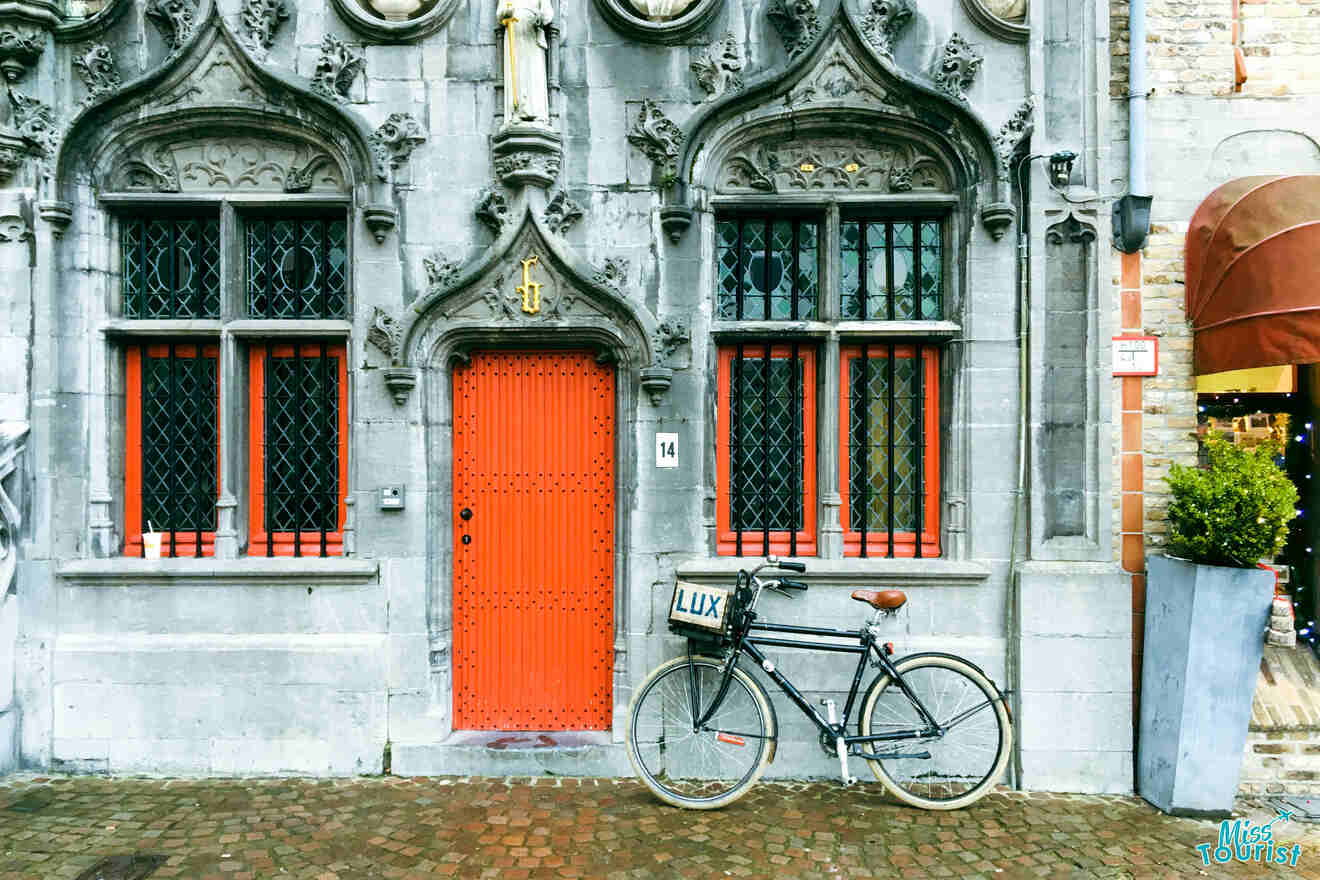 Vintage bicycle parked in front of a historic grey stone building with ornate windows and a bright orange door in Bruges