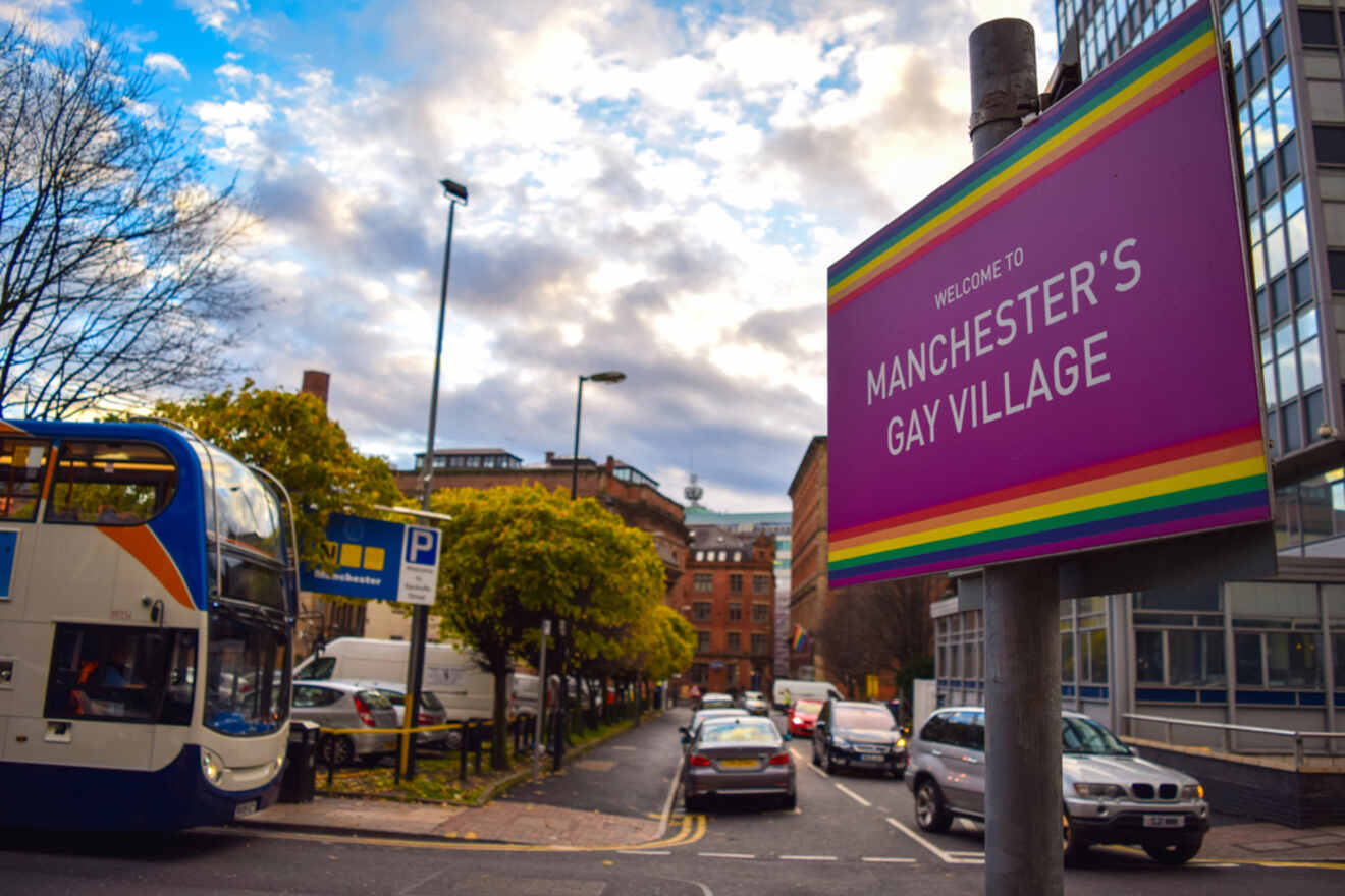 A colorful welcome sign for "Manchester's Gay Village" with a rainbow motif, with buses and cars on the street indicating a lively city area.