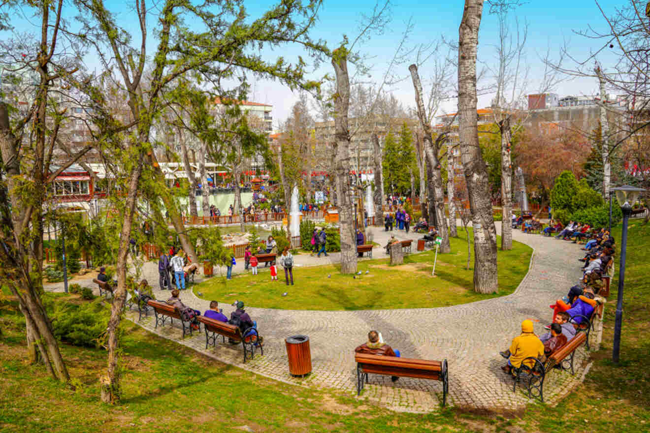 Vibrant scene at Kuğulu Park in Ankara, where locals enjoy leisure time sitting on benches, walking, and socializing in a serene park setting with tall trees and colorful flowers.