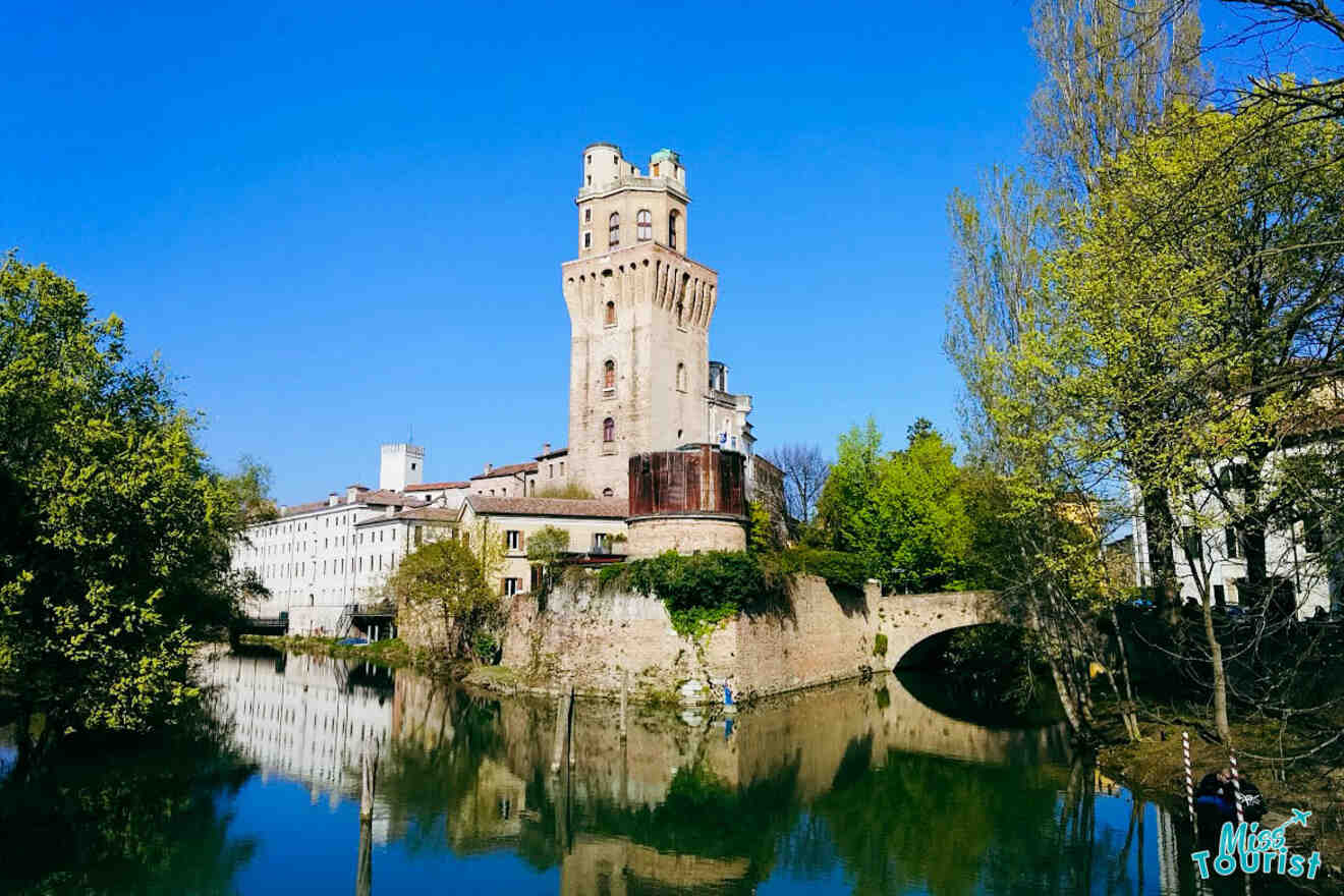 Specola Observatory tower in Padua reflecting on a calm river surrounded by lush greenery and a clear blue sky