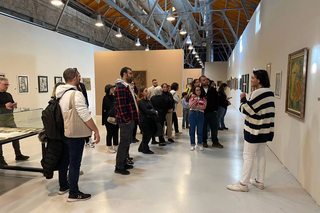 Visitors touring the Cermodern art gallery, attentively listening to a guide in a spacious hall adorned with a variety of framed artworks on the walls and display cases with documents