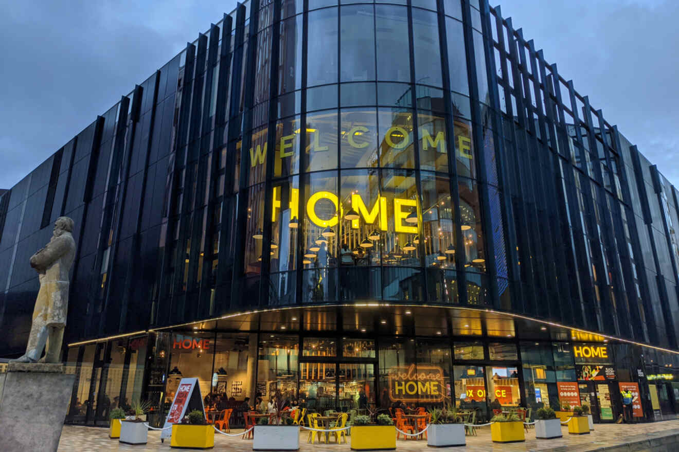 Facade of HOME theater in Manchester at dusk, featuring large glass windows with bold yellow 'WELCOME HOME' letters, and a statue to the left.