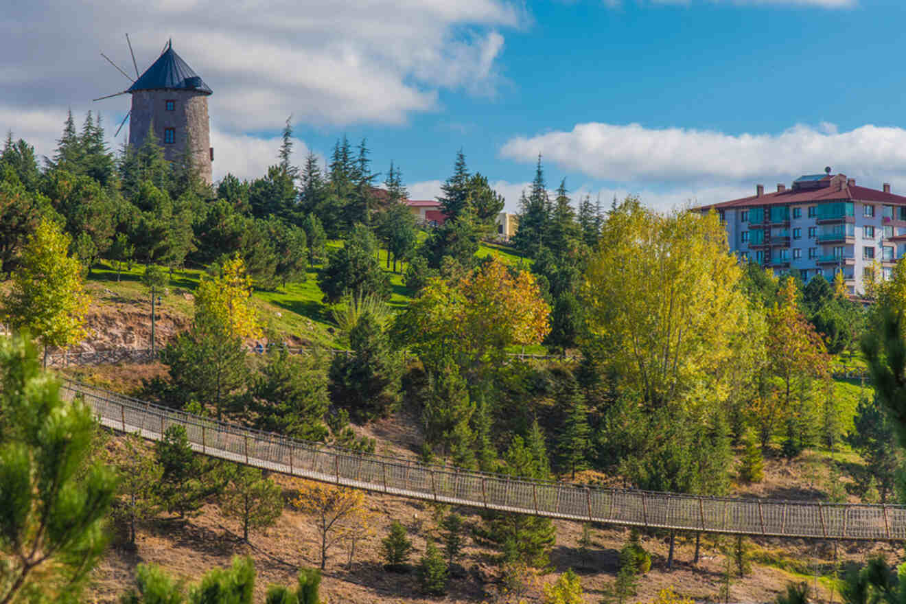 A picturesque view of Altınköy Open Air Museum in Ankara, with a historic windmill atop a lush green hill and a suspended bridge amidst the trees, under a blue sky with fluffy clouds.