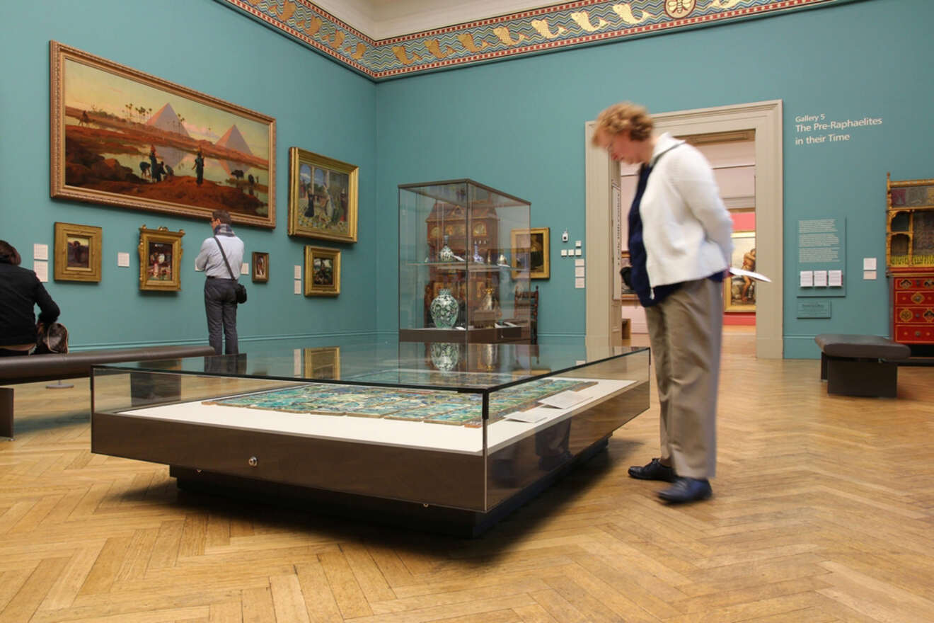 Visitors observing artworks at the Manchester Art Gallery, featuring teal walls, classical paintings, and a display case in the center of the room